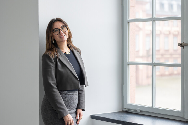 Porträtbild von Joana Furhmann. Sie hat halblange braune Haare und eine Brille, trägt einen grauen Blazer und einen grauen Rock und steht neben einem Fenster.