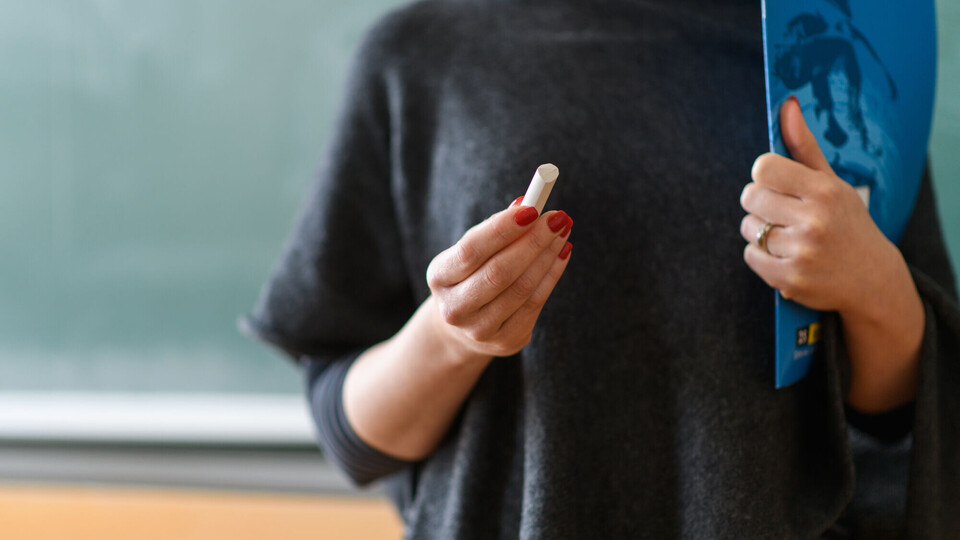 Eine Frau im schwarzen T-Shirt steht vor einer grünen Kreidetafel und hält ein stück Kreide in der manikürten Hand und ein Heft unterm Arm. Eine Lehrerin?