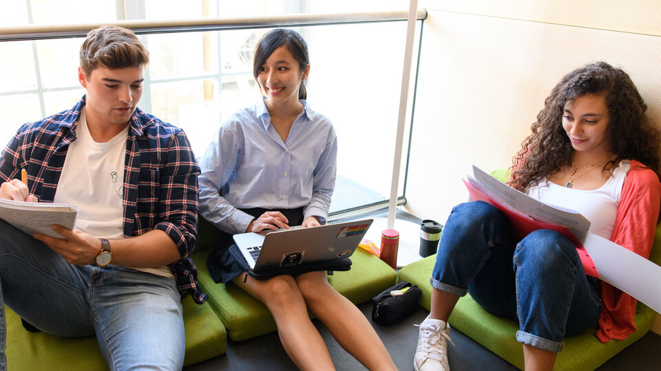 Drei Studierende sitzen nebeneinander auf grünen Sitzkissen mit ihren Laptops oder Schreibblöcken.