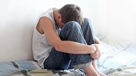 Ein Teenager sitzt mit angezogenen Beinen auf seinem Bett. Sein Kopf ist auf den Knien abgelegt und die Arme um seine Beine geschlungen.